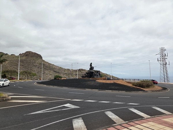 Rotonda en Adeje con escultura del mencey guanche frente al mar, con laderas volcánicas al fondo en un día nublado.