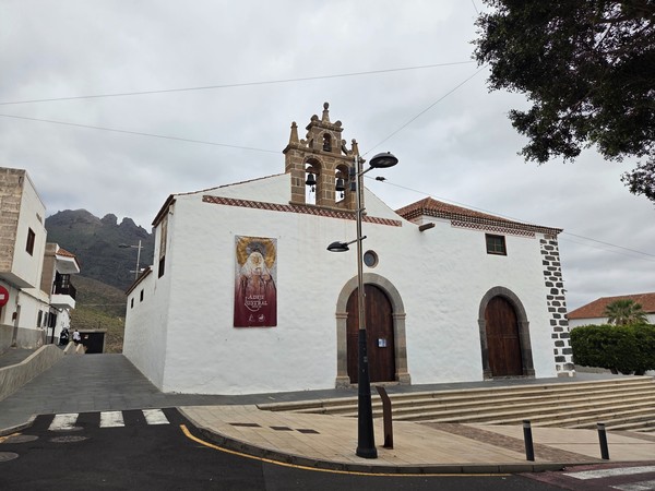 Fachada encalada de la Iglesia de Santa Úrsula en Adeje, con campanario de piedra, escalinata y montañas al fondo en un día nublado.