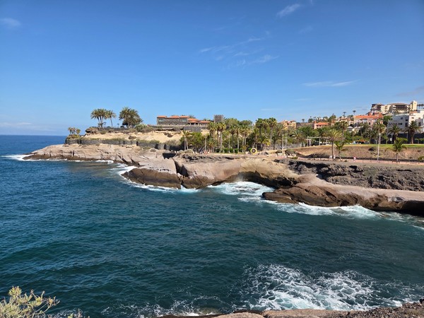 Vista de Costa Adeje con acantilados de lava, palmeras y paseo marítimo sobre un mar azul intenso.