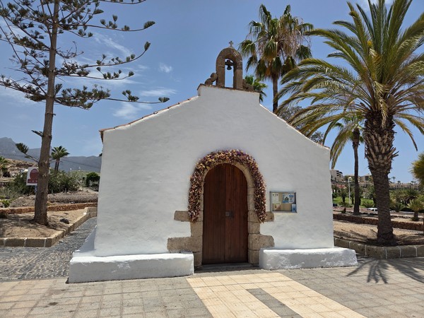Pequeña ermita encalada en Costa Adeje con puerta de madera y arco floral, flanqueada por palmeras bajo cielo azul.