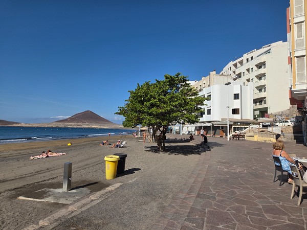 Playa urbana de El Médano con bañistas, paseo peatonal arbolado y la silueta volcánica de Montaña Roja frente al mar.