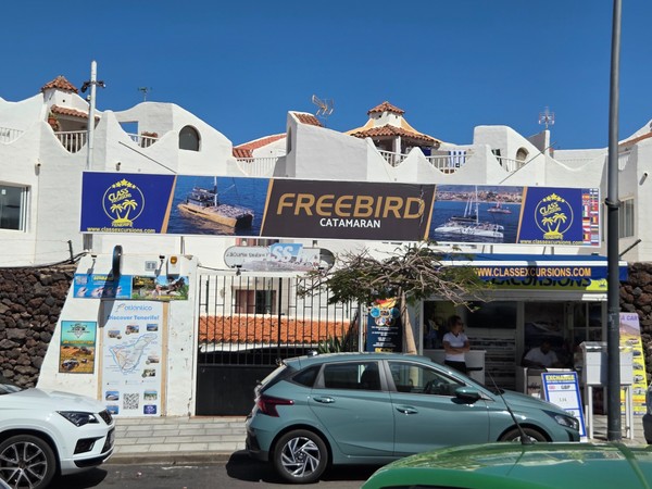 Fachada de la oficina de Freebird Catamaran con carteles de excursiones marítimas y coches aparcados delante.