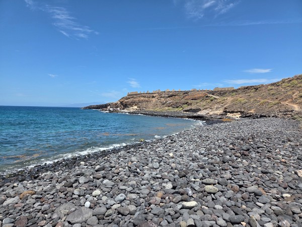 Orilla de cantos rodados negros y aguas azules en la Playa de Los Morteros, con laderas áridas y acantilado al fondo.